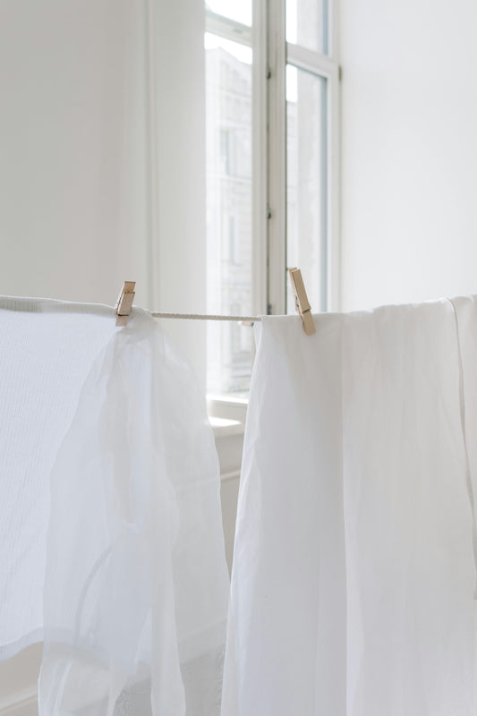 Two white linen panels hang gently from drying lines, illuminated by soft daylight streaming through a window—evoking natural care and timeless textile rituals.