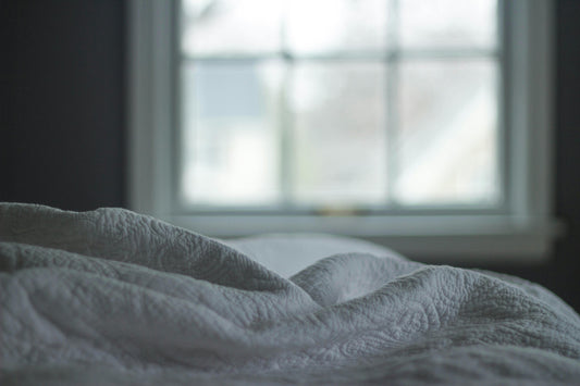 White linen blanket draped across a bed, softly lit by morning sunlight streaming through a window.