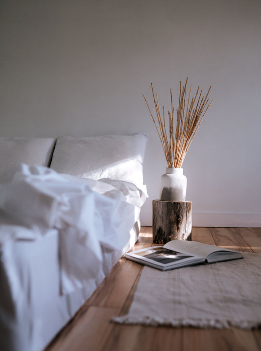 White linen quilt on bed with minimal design. Wooden floors and open book atop a rug
