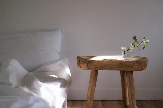 Sunlight streaming across a bed with white linen and pillows, beside a wooden side table topped with a single flower.