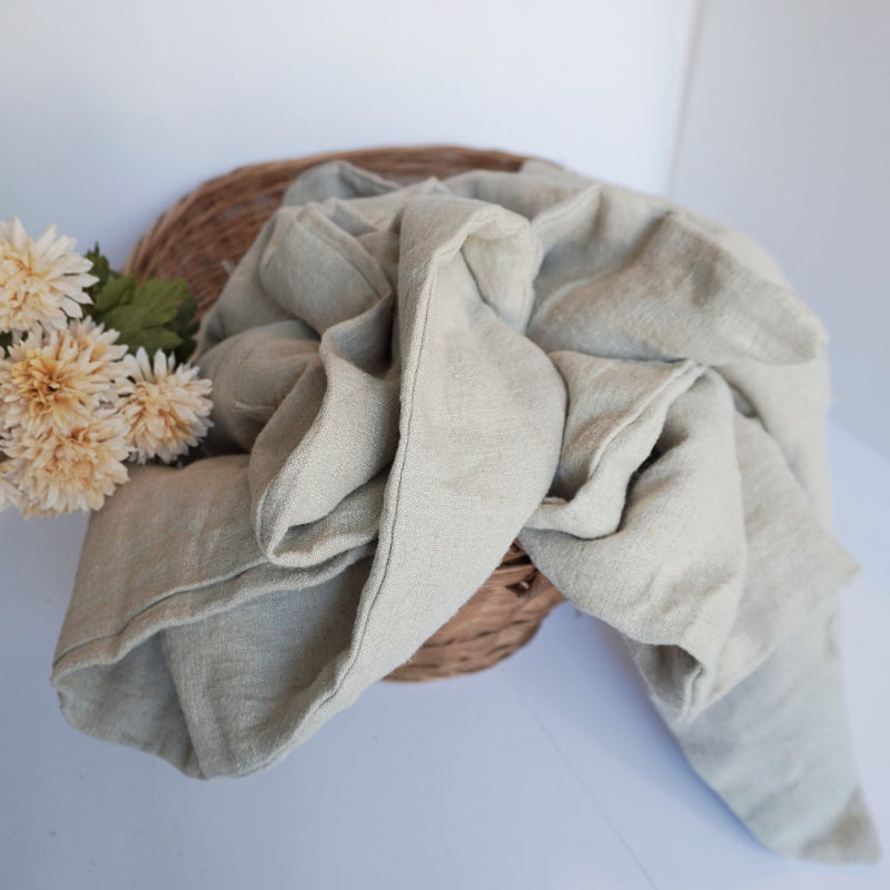 A light sage green linen throw blanket draped over a woven wicker basket with cream-colored flowers, set against a clean white background.