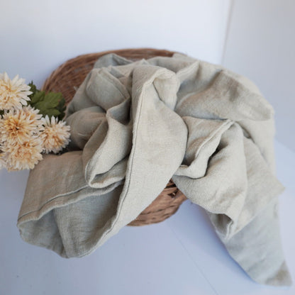 A light sage green linen throw blanket draped over a woven wicker basket with cream-colored flowers, set against a clean white background.