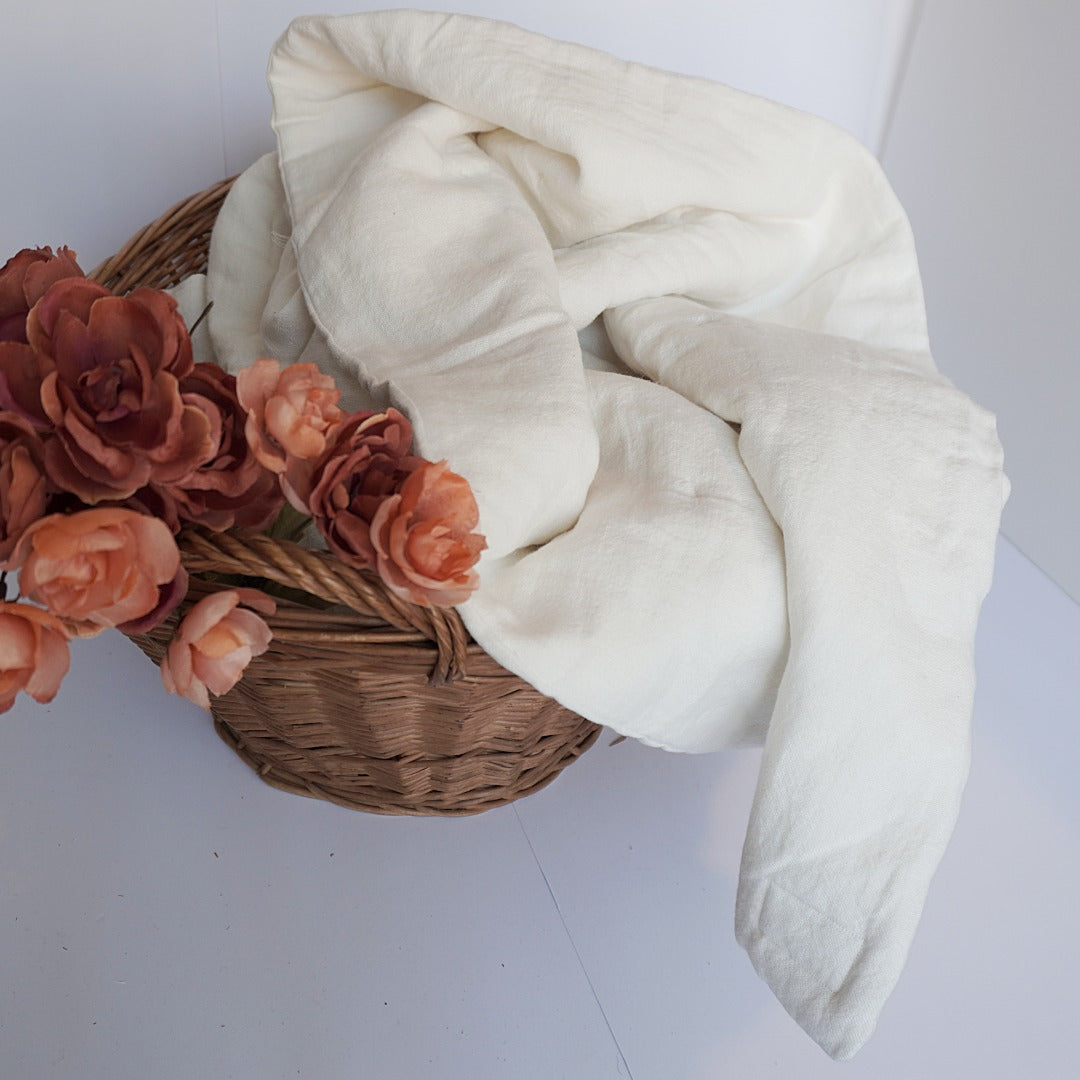 An off-white, textured linen blanket draped over a brown woven basket filled with peach-colored artificial roses, set against a plain white background.