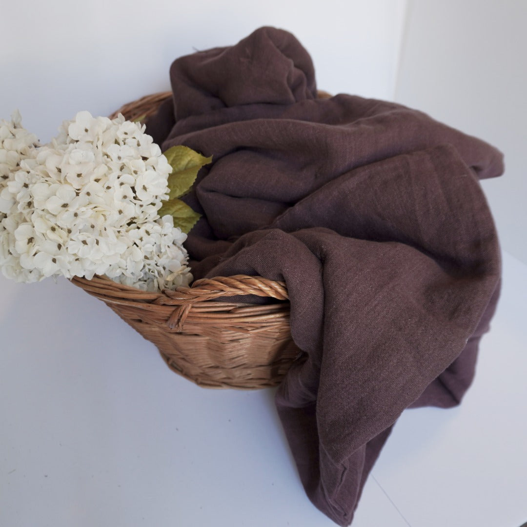 A draped dark brown linen quilt or throw blanket, styled in a wicker basket with white hydrangea flowers on a plain white background.