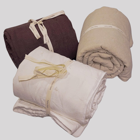 Three rolled bedding items, likely blankets or sheets, in dark brown, cream, and light beige, each tied with a cream ribbon. The cream ribbon has small printed text that appears to read 'SUNRISE LINEN'.