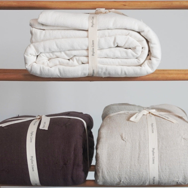Three folded linen blankets in cream, dark brown, and light gray, each tied with a cream ribbon printed with the brand name 'Peplos Linen', displayed on wooden shelves.