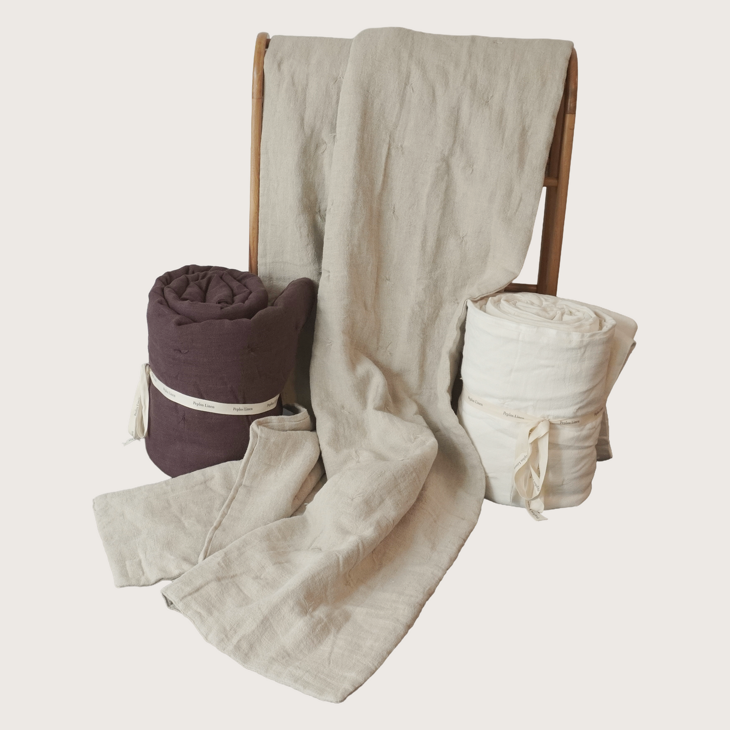 Three rolled and draped textile linen blankets or quilts on a wooden stand against a plain light background: a large cream linen-like cloth draped over the center, a rolled cream piece on the right, and a rolled deep plum-purple piece on the left with a white band.