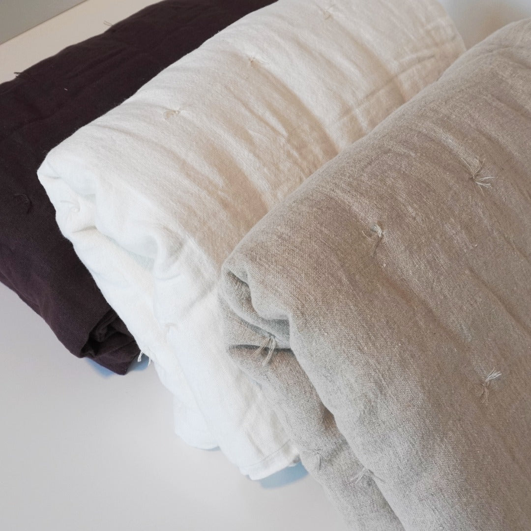 Three folded textured linen quilts in dark brown, off-white, and light gray-beige, arranged diagonally on a white surface.