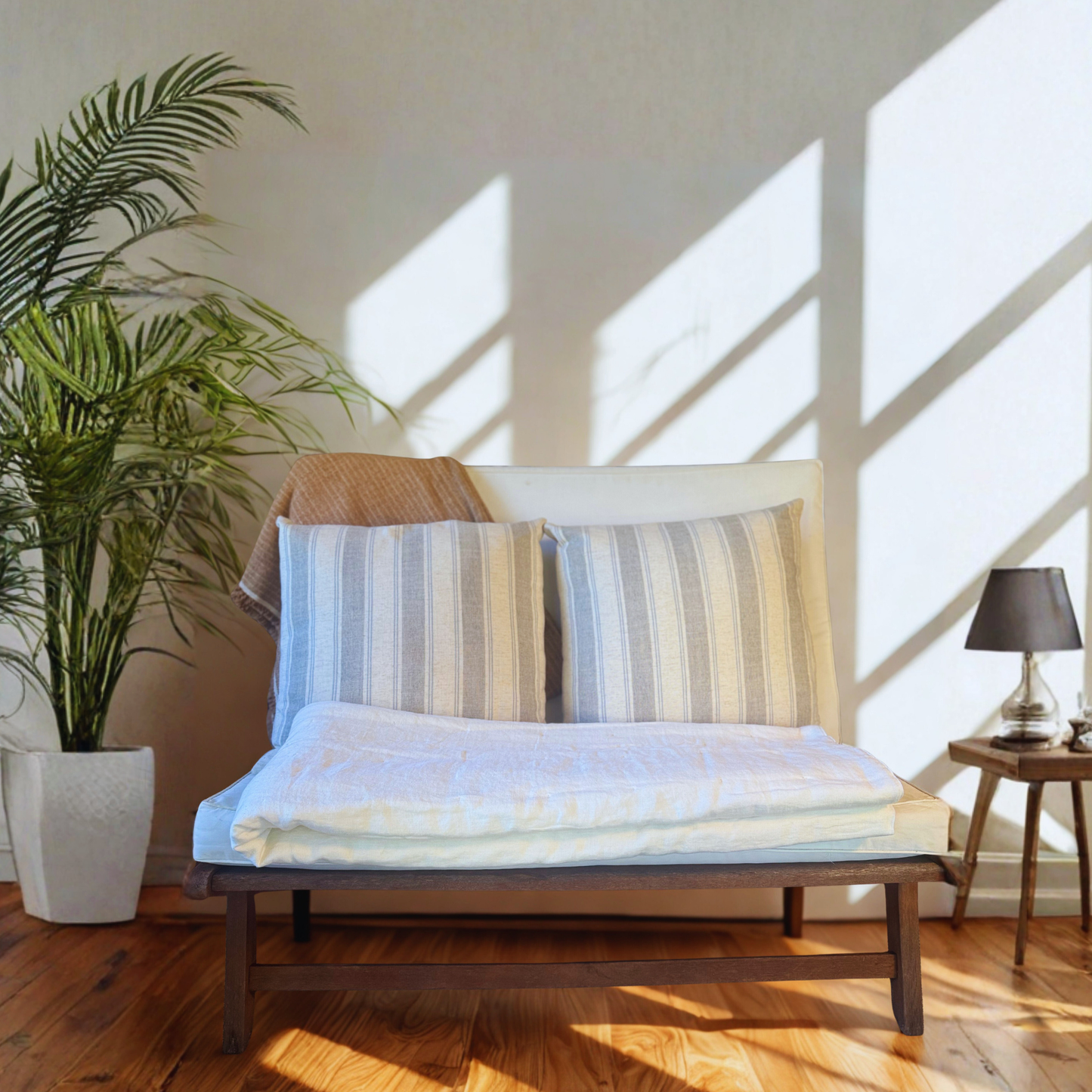 Bench with striped pillows in a bright room with sunlight streaming through a large window and a folded ivory linen quilt