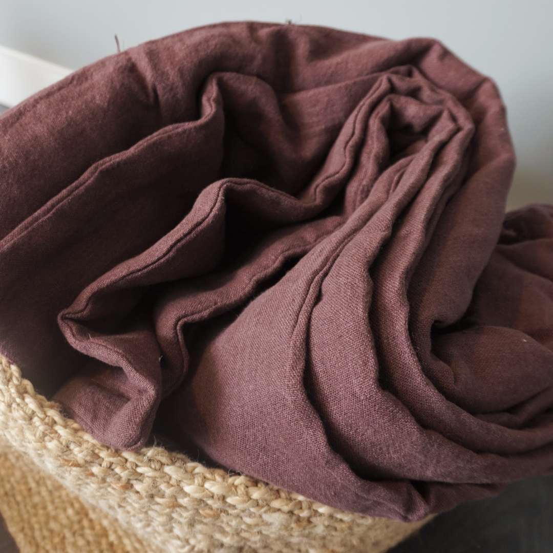 A close-up of a folded brown or mauve-colored linen quilt, likely a throw blanket inside a woven jute basket.