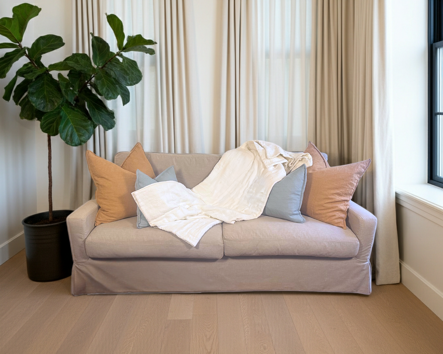 Beige sofa with colorful pillows and a linen blanket in a room with wooden flooring and a plant.