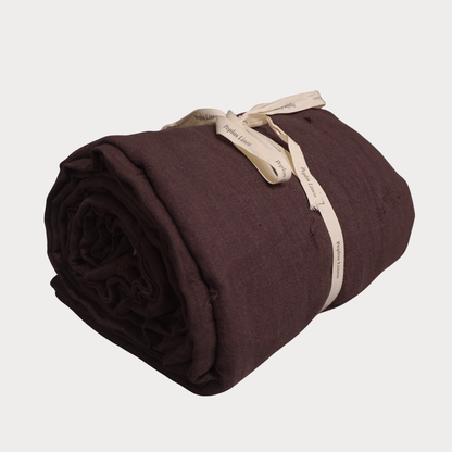 A rolled dark brown linen blanket tied with a cream-colored paper ribbon, shown on a light gray background.