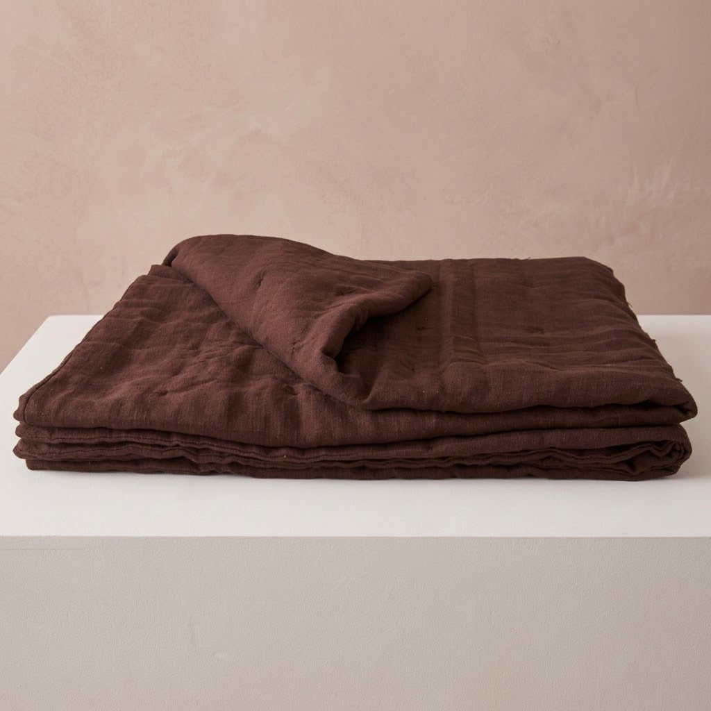 Chocolate brown linen quilt folded on a white surface, showing its soft, textured layers and relaxed drape.