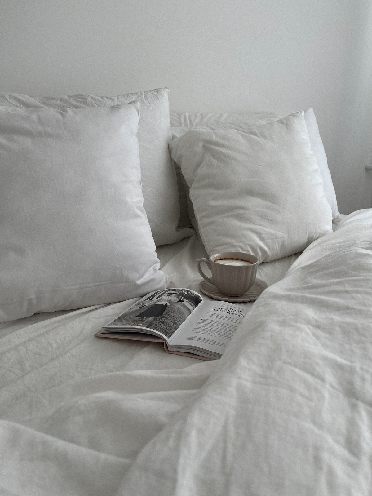 White pillows and linen blanket with a book and cup on a bed